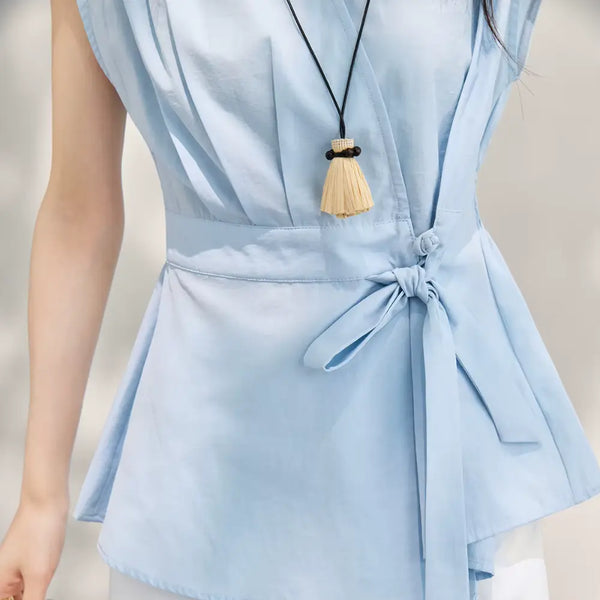 Light blue sleeveless top with a tie waist and a necklace with a tassel on a neutral background