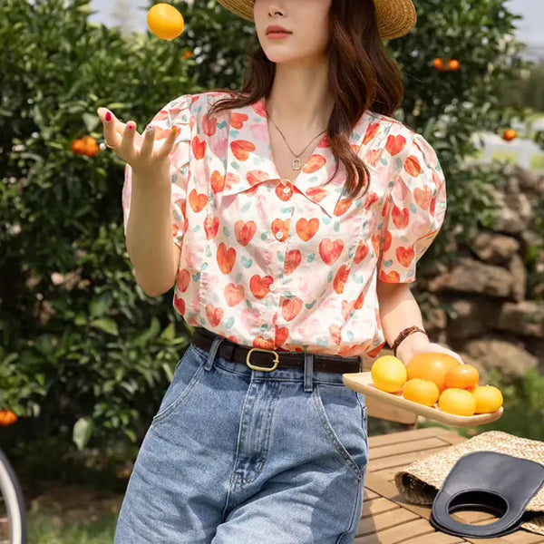 Woman in a heart-patterned shirt and straw hat holding oranges in an orchard.