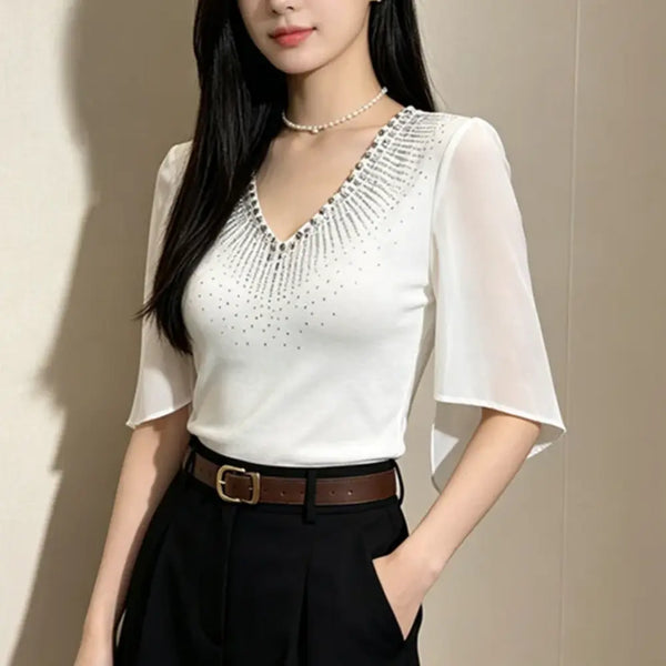 A woman wearing a white short-sleeve V-neck blouse with a rhinestone embellished yoke, paired with black trousers and a brown belt, standing against a neutral beige wall.
