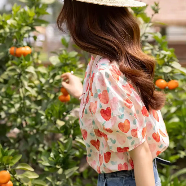 Woman wearing a heart-patterned blouse in an outdoor setting with orange fruits and green foliage.