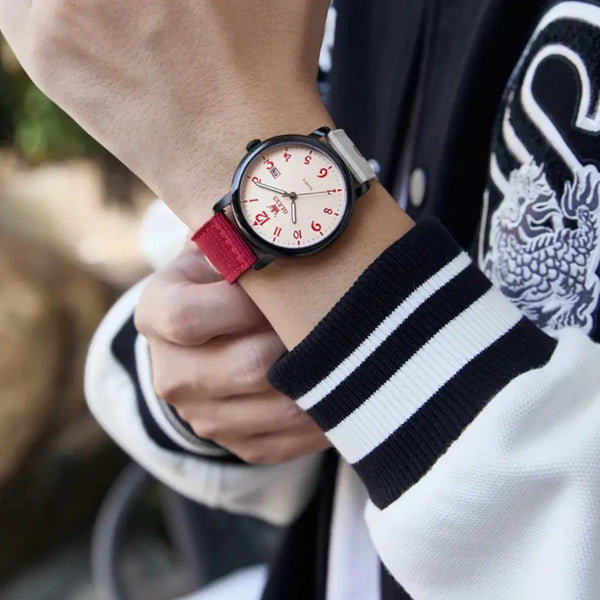 Person wearing a watch with a red strap and white face, holding a blurred outdoor background.