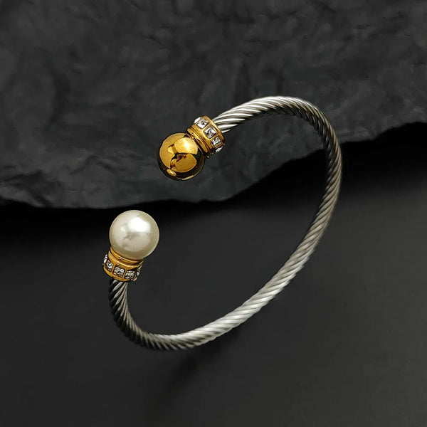 A twisted cable-style cuff bracelet in silver-tone metal with one end featuring a white pearl and the other a polished gold-tone ball charm, both accented with small clear stones, displayed on a dark textured surface.