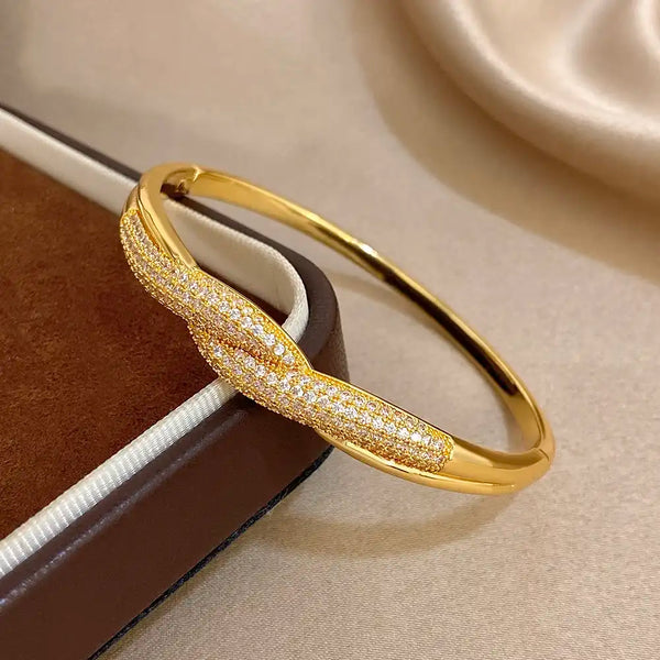 A gold hinged bangle bracelet with a twisted knot design, featuring pavé-set clear stones on one half and a polished gold finish on the other, resting on a brown jewelry box.