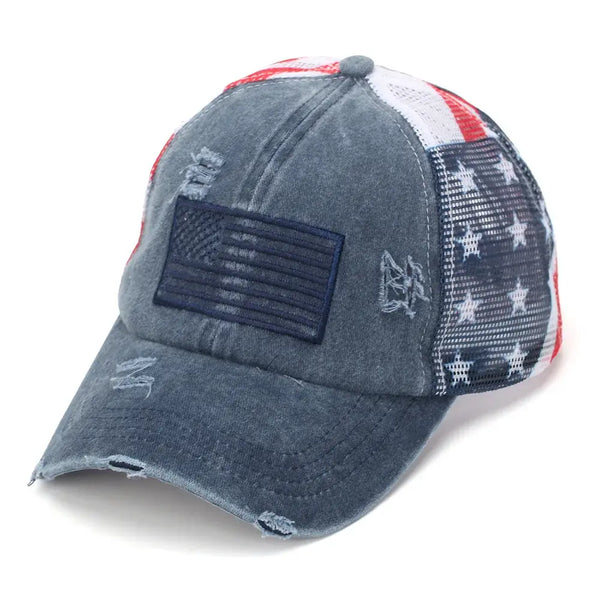 A distressed trucker-style baseball cap featuring a washed blue-gray front and brim, a navy American flag patch on the front, and a red, white, and blue mesh back with white stars.