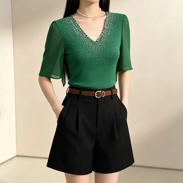 A woman wearing a green short-sleeve V-neck blouse with rhinestone embellishments around the neckline, paired with black high-waisted shorts and a brown belt, standing against a plain indoor wall.