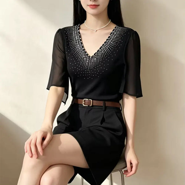 A woman seated indoors wearing a black sheer short-sleeve V-neck top with sparkling embellishment around the neckline, paired with a black skirt and brown belt.
