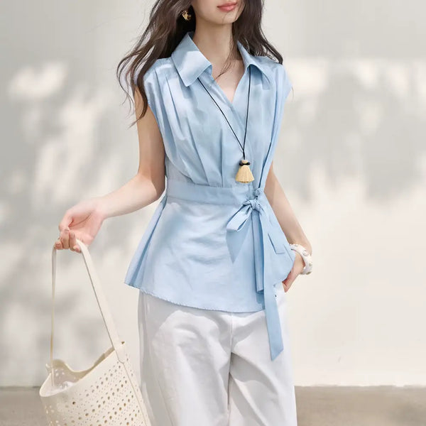 Woman wearing a light blue sleeveless blouse with a tie waist, holding a beige handbag against a neutral background.