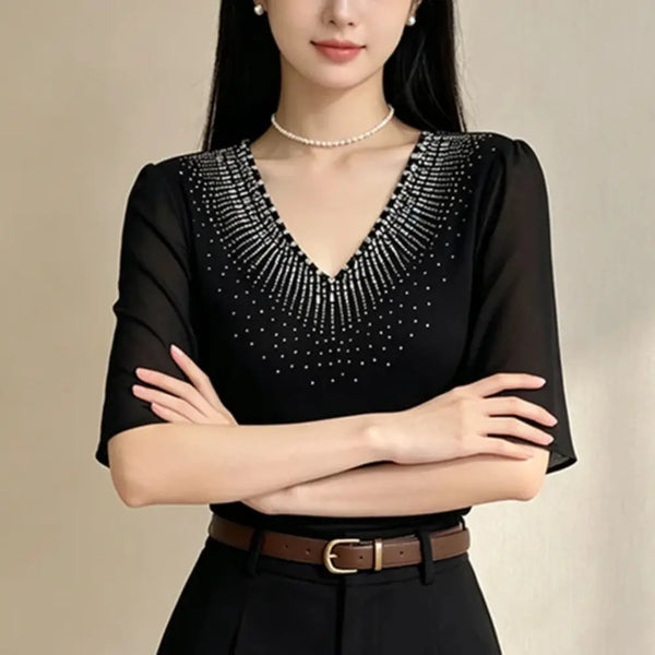 A woman wearing a black short-sleeve V-neck blouse with sparkling silver rhinestone embellishments around the neckline, paired with a brown belt and black skirt against a neutral beige background.
