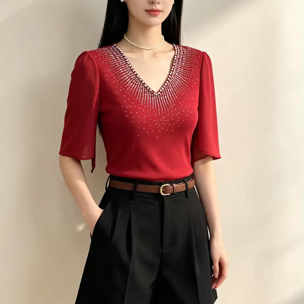 A woman wearing a red short-sleeve V-neck blouse with sparkling embellishments around the neckline, paired with black high-waisted shorts and a brown belt, standing against a plain light beige wall.