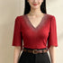 A woman wearing a red short-sleeve V-neck blouse with sparkling rhinestone embellishments around the neckline, paired with a brown belt and black skirt against a plain beige background.