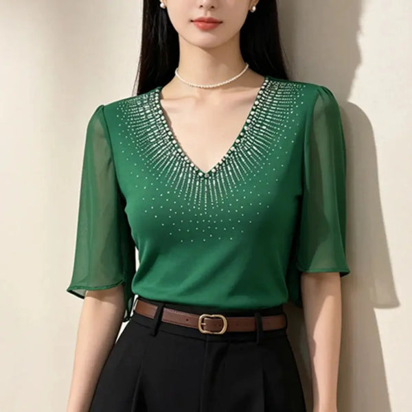 A woman wearing a green short-sleeve V-neck blouse with a sparkling rhinestone embellished neckline, paired with black trousers and a brown belt, standing against a light beige wall.