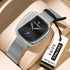 Silver watch with black face on a beige surface, featuring a visible tag with price and brand name.
