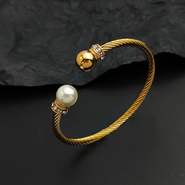 A gold-toned twisted cable open bangle bracelet featuring a white pearl and small clear stones, displayed on a dark textured background.
