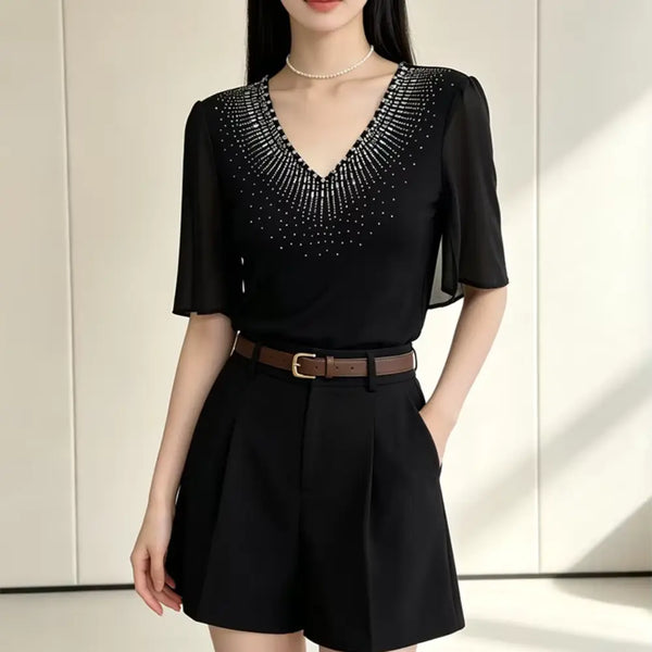 A woman wearing a black short-sleeve blouse with a V-neckline and sparkling embellished yoke, paired with black pleated shorts and a brown belt, standing against a light beige paneled wall.