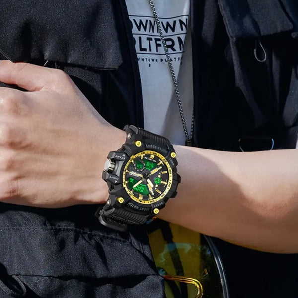 A close-up of a person's wrist wearing a rugged black sports watch with a green dial and gold accents, paired with a black jacket and white graphic t-shirt.