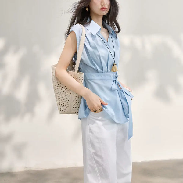 Woman wearing a light blue sleeveless top and white pants, holding a woven bag against a neutral background.