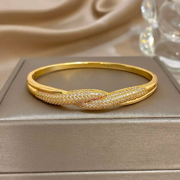 A gold hinged bangle bracelet with a twisted knot design, featuring one side in polished gold and the other in pavé-set clear stones, displayed on a textured gray jewelry box.