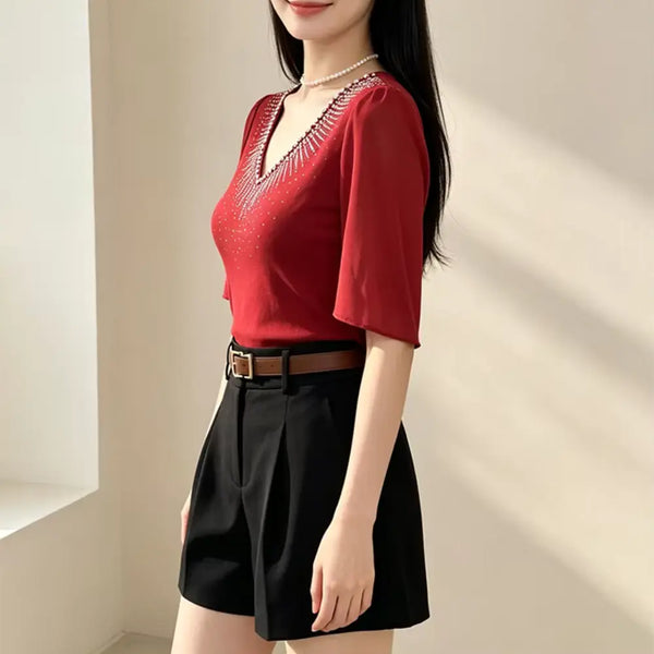 A woman wearing a red short-sleeve V-neck top with a decorative silver embellished neckline, paired with black high-waisted shorts and a brown belt, standing in a brightly lit indoor setting.