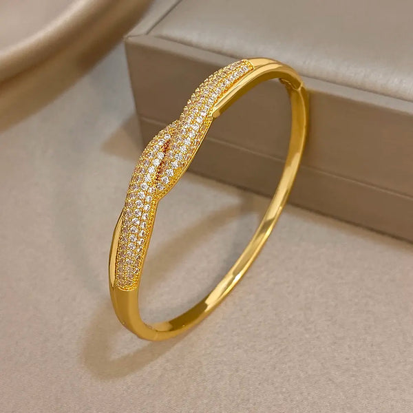 A polished gold bangle bracelet featuring a central twisted knot design paved with small clear stones, displayed on a neutral beige surface next to a jewelry box.