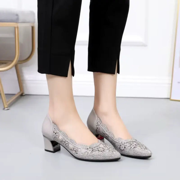 Gray high-heeled shoes with intricate designs worn by a person in black pants on a light gray floor.